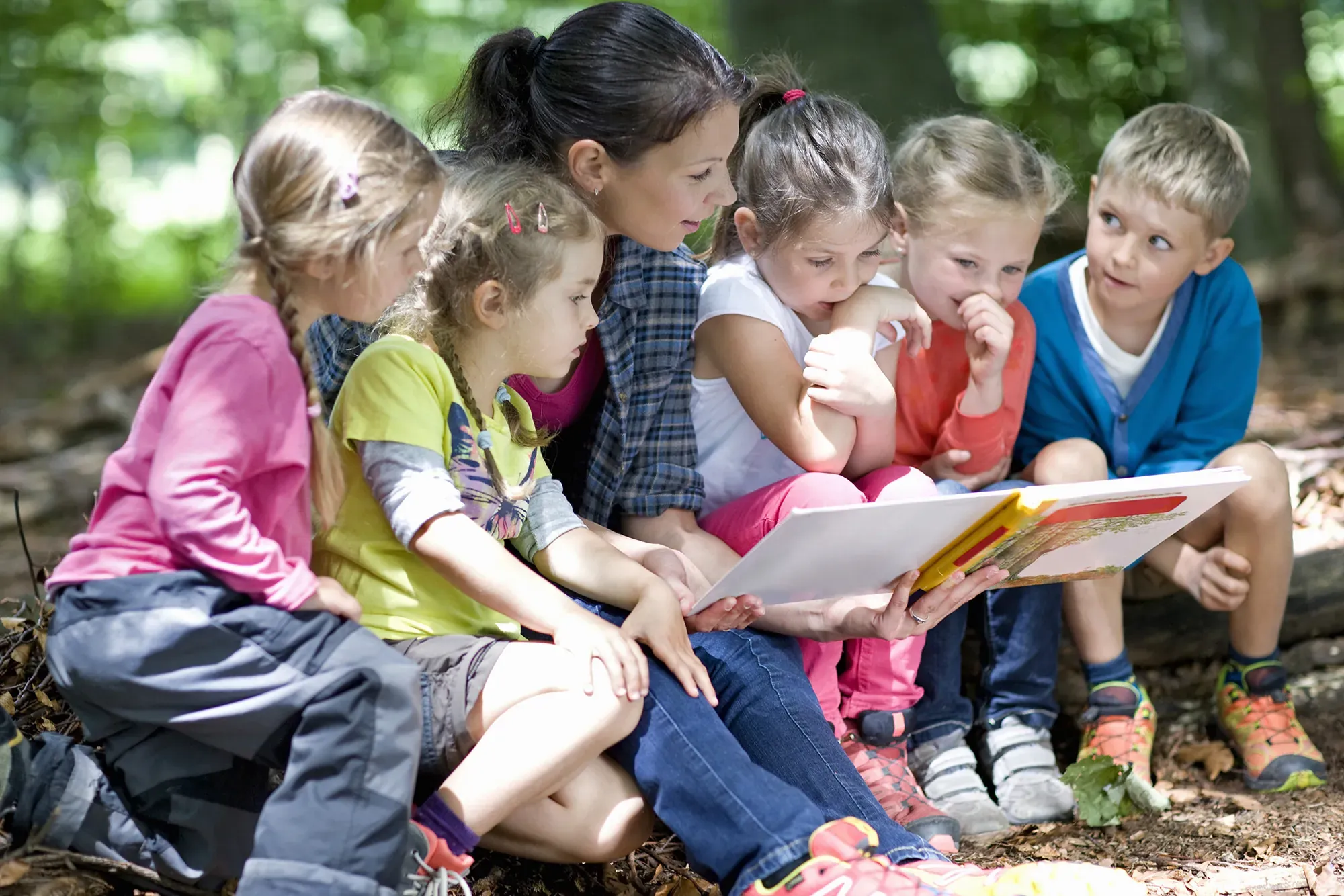 An adult reading a picture book to five young children sitting together in woodland