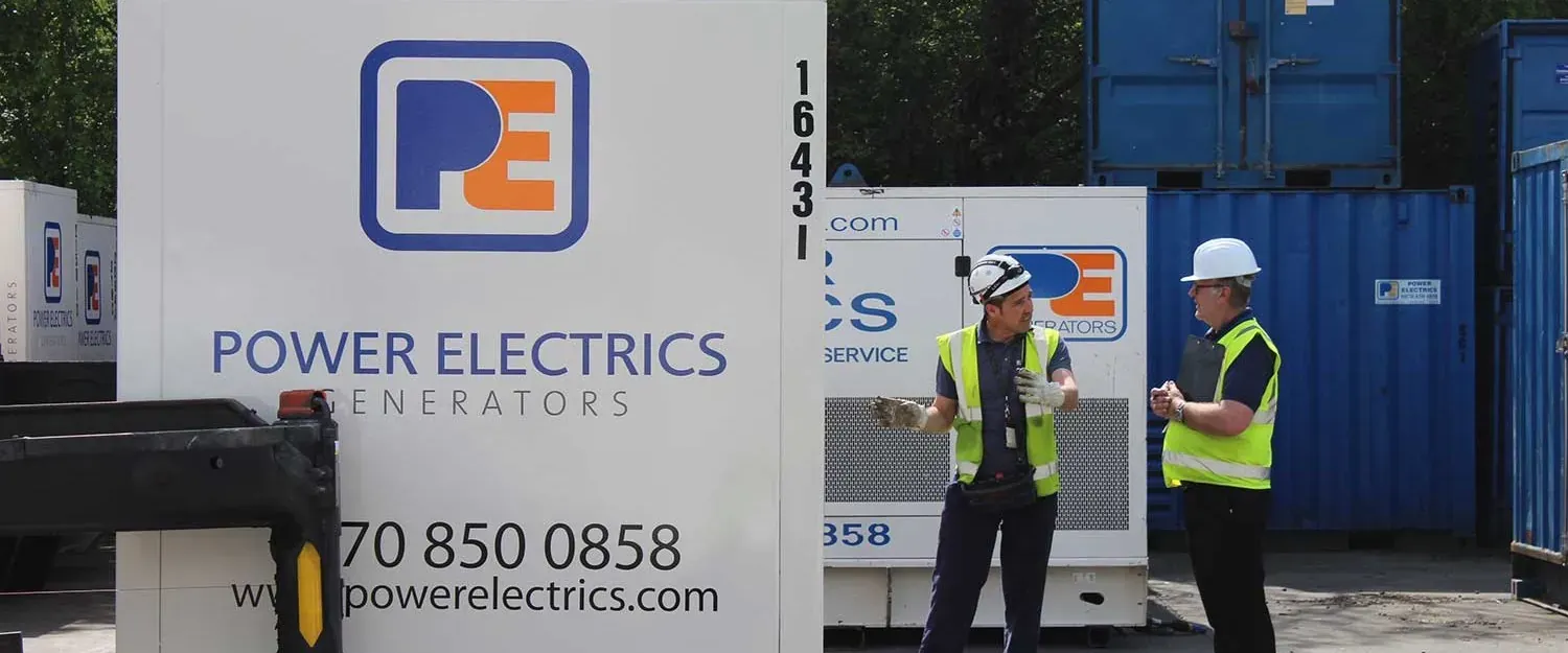 Two men in hard hats and high-visibility vests talking beside Power Electrics generators in a yard